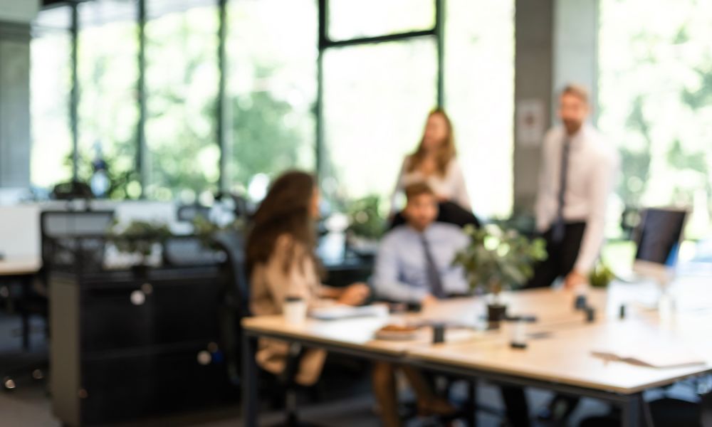 Blurred image of a modern office with four people having a discussion near desks and greenery.