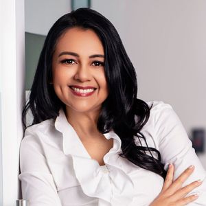 Margarita Eberline with long black hair wearing a white blouse, standing confidently indoors.