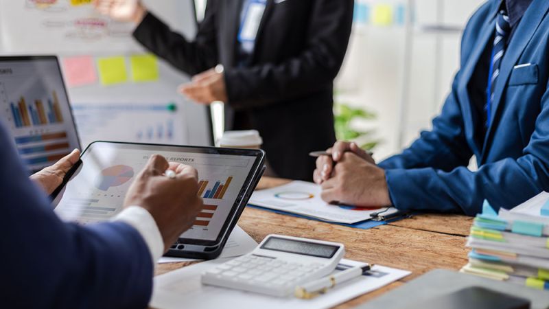 Business team analyzing charts on tablet and documents during a meeting, with a whiteboard in the background.