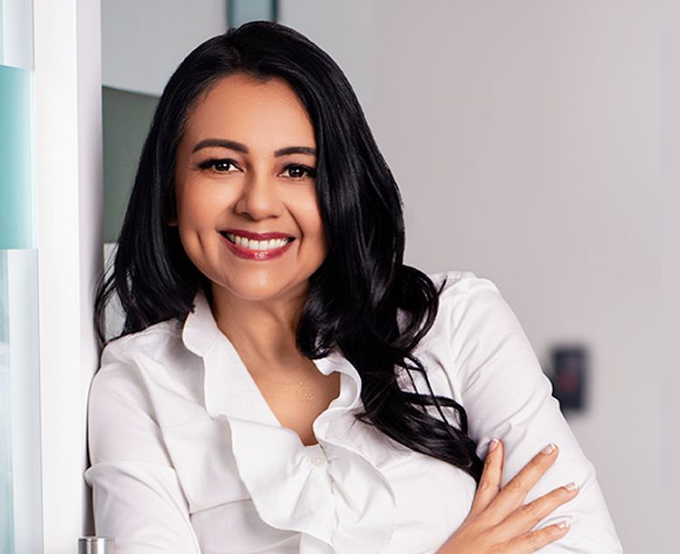 Smiling woman in white blouse, leaning against a wall, in a modern office setting.